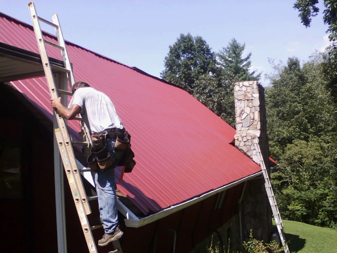 Roofer on a ladder beside a red metal roof and stone chimney — Soffit & Fascia Repair by Roofing Brothers in Saulsbury